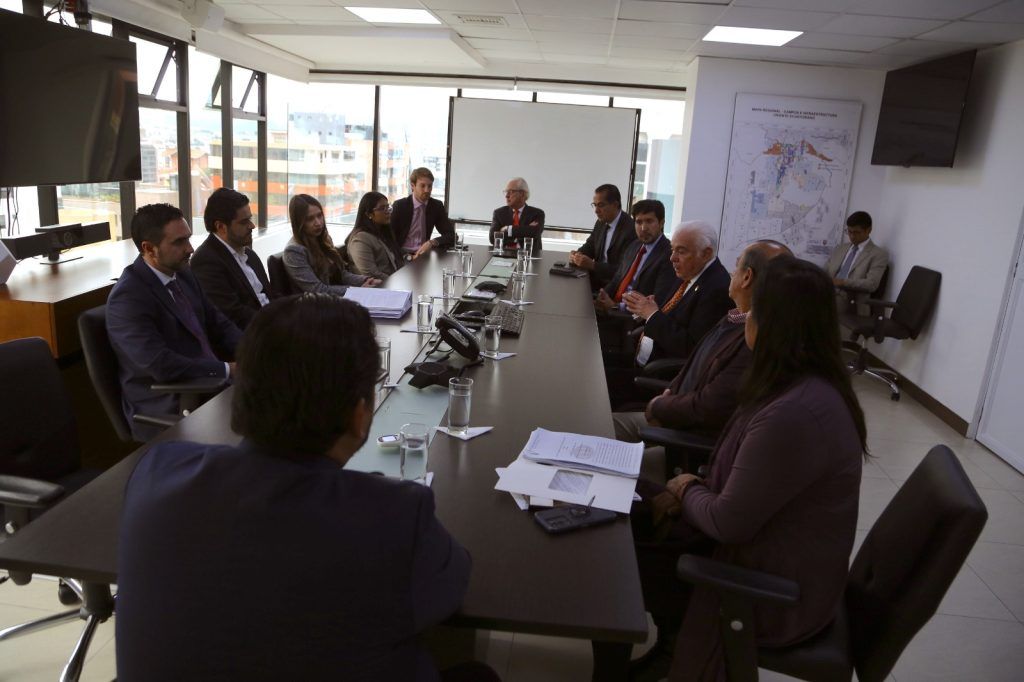 Grupo de profesionales en trajes de negocios participando en una reunión en una oficina con mapas y pantallas de presentación al fondo.